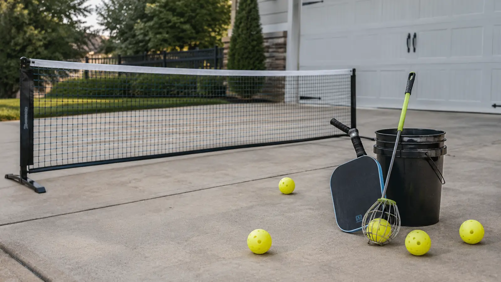 a portable pickleball net in a driveway with balls on the ground and a paddle with a retriever nearby
