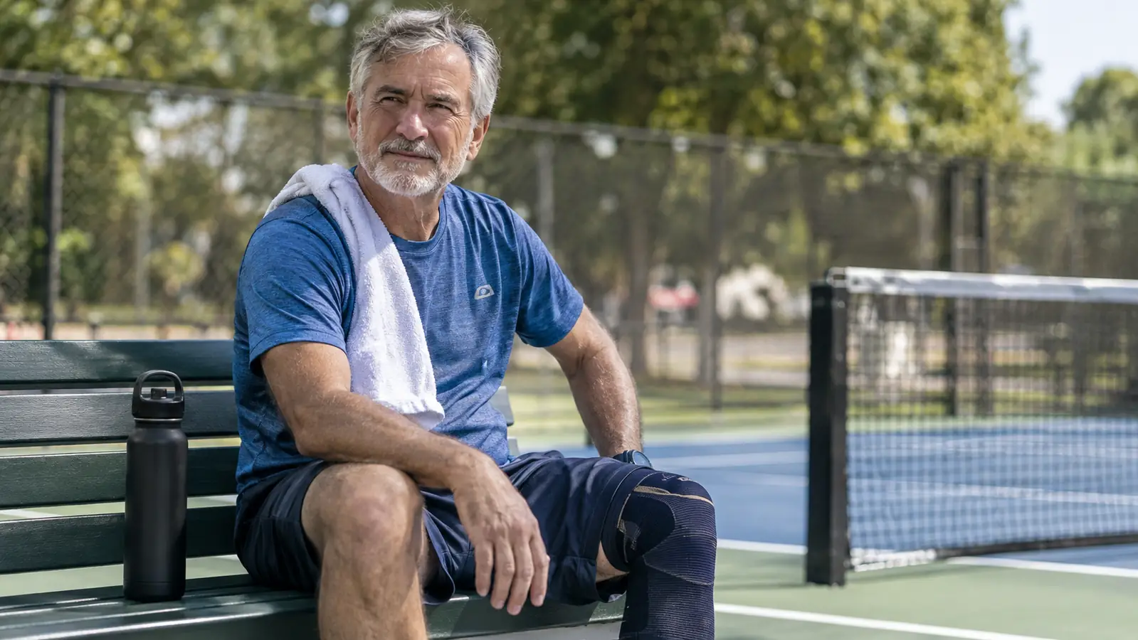 A man in his sixties sitting on a bench after a game, one hand on his knee, with a compression sleeve visible below his shorts