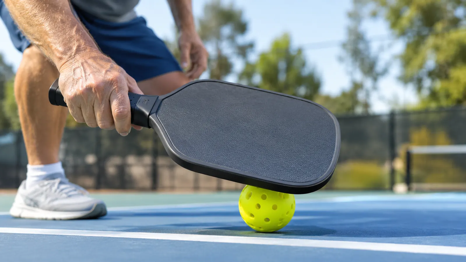 A hand using a paddle-mounted ball retriever to pick up a pickleball from the court surface