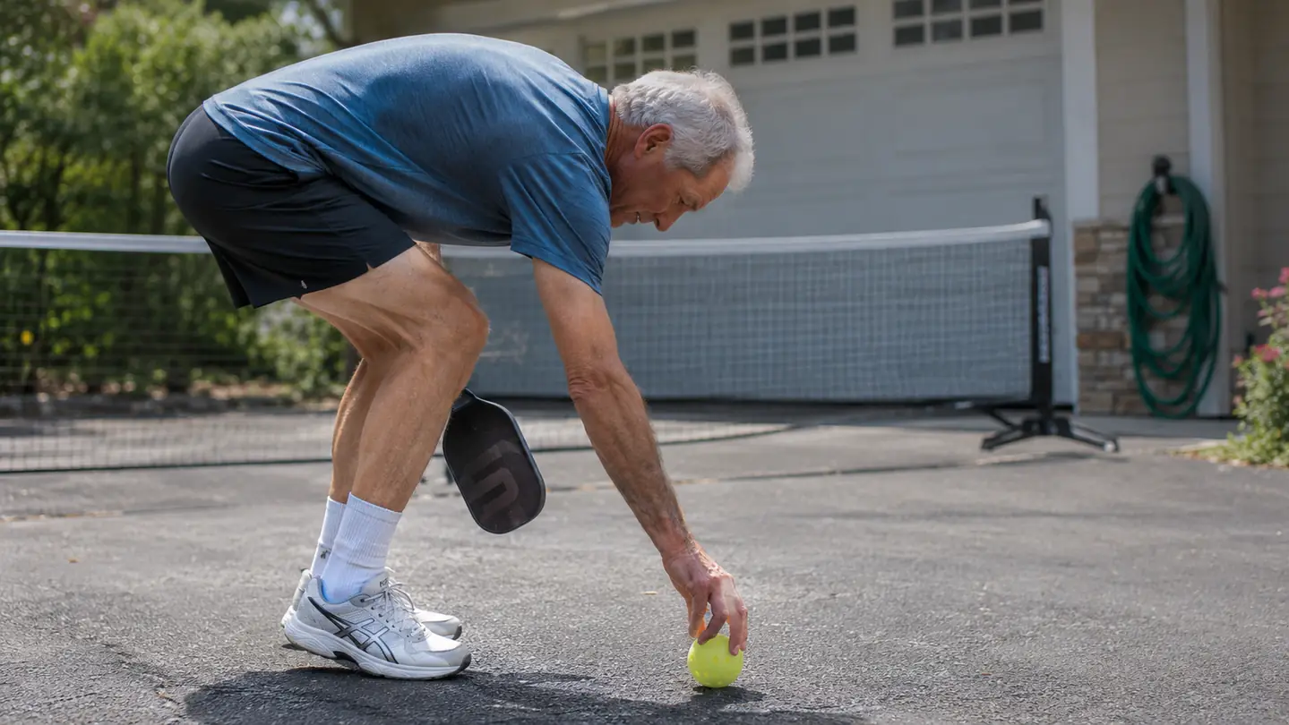 A man in his sixties bending to pick up a pickleball from the driveway, the portable net behind him